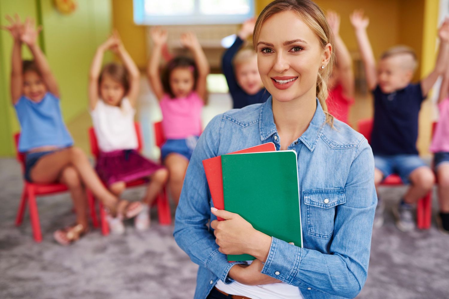 A smiling teacher holding folders in front of a blurred background of students with their hands raised.