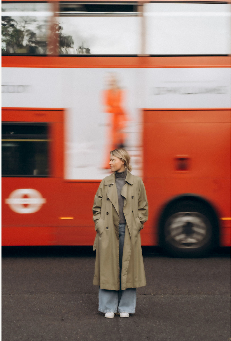 Woman in beige trench coat standing against blurred red London bus background.