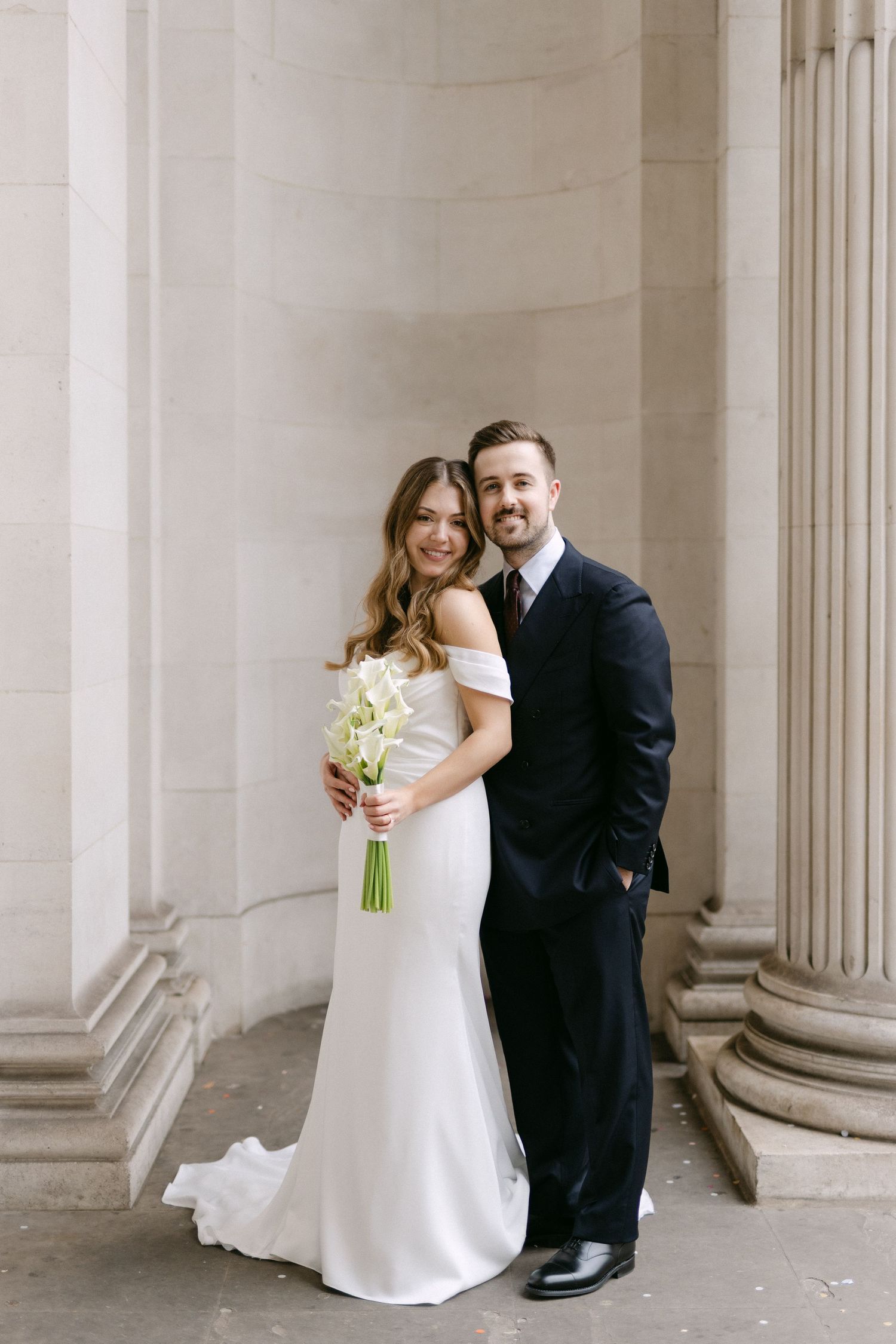 Bride in white off-shoulder gown holding calla lily bouquet poses with groom in black suit beside stone columns.