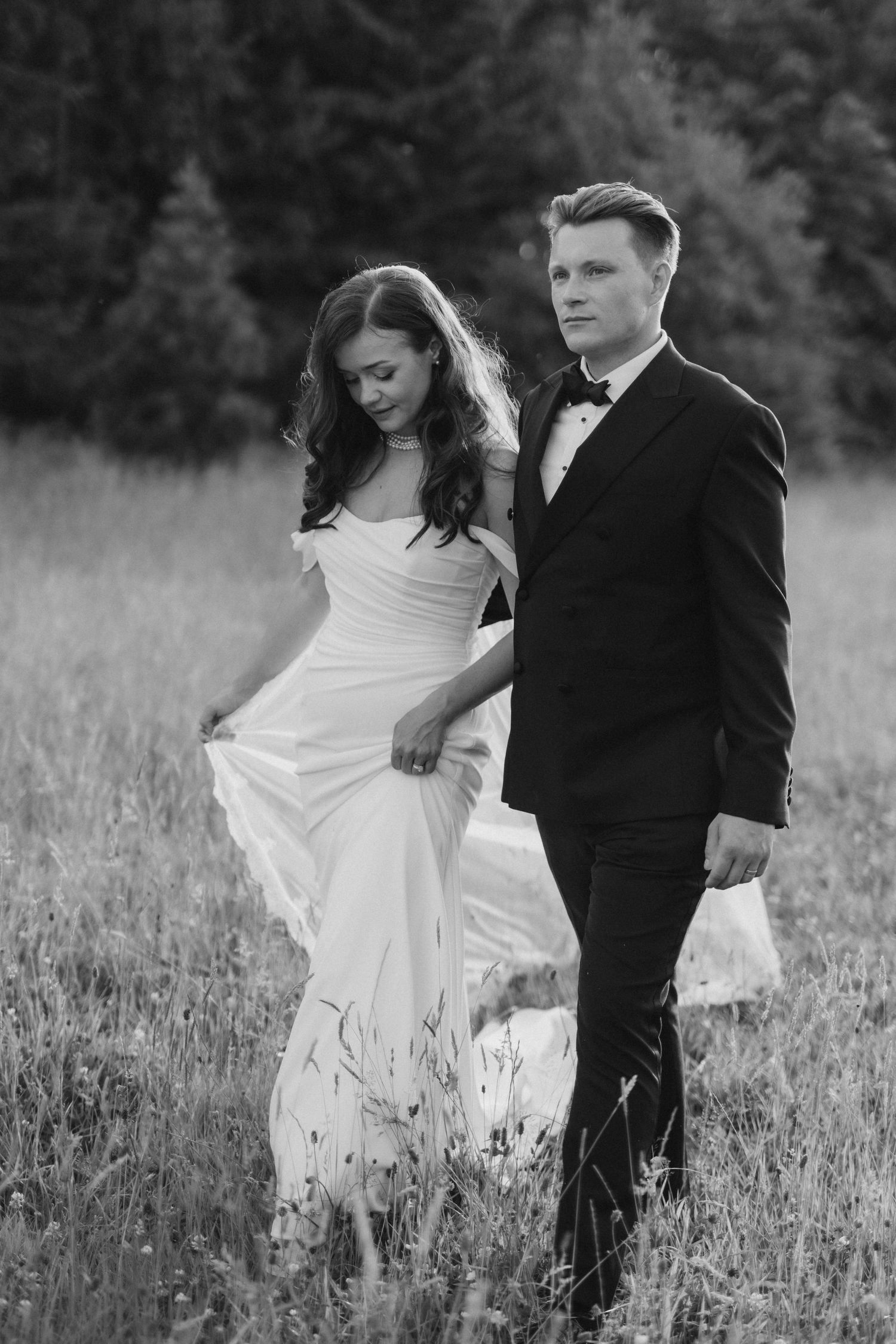 Black and white photo of a bride and groom walking in a meadow, bride in flowing gown, groom in tuxedo.