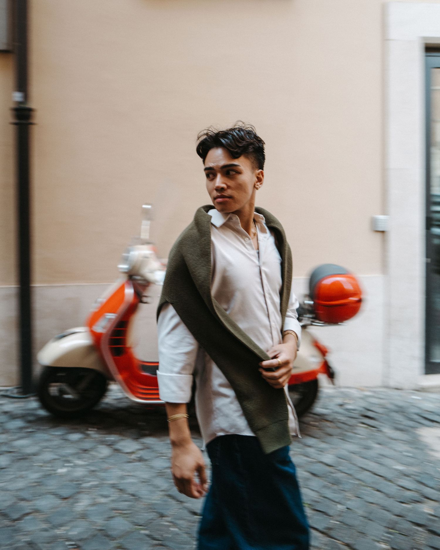 A street style photo with a red Vespa scooter in the background on cobblestone streets.