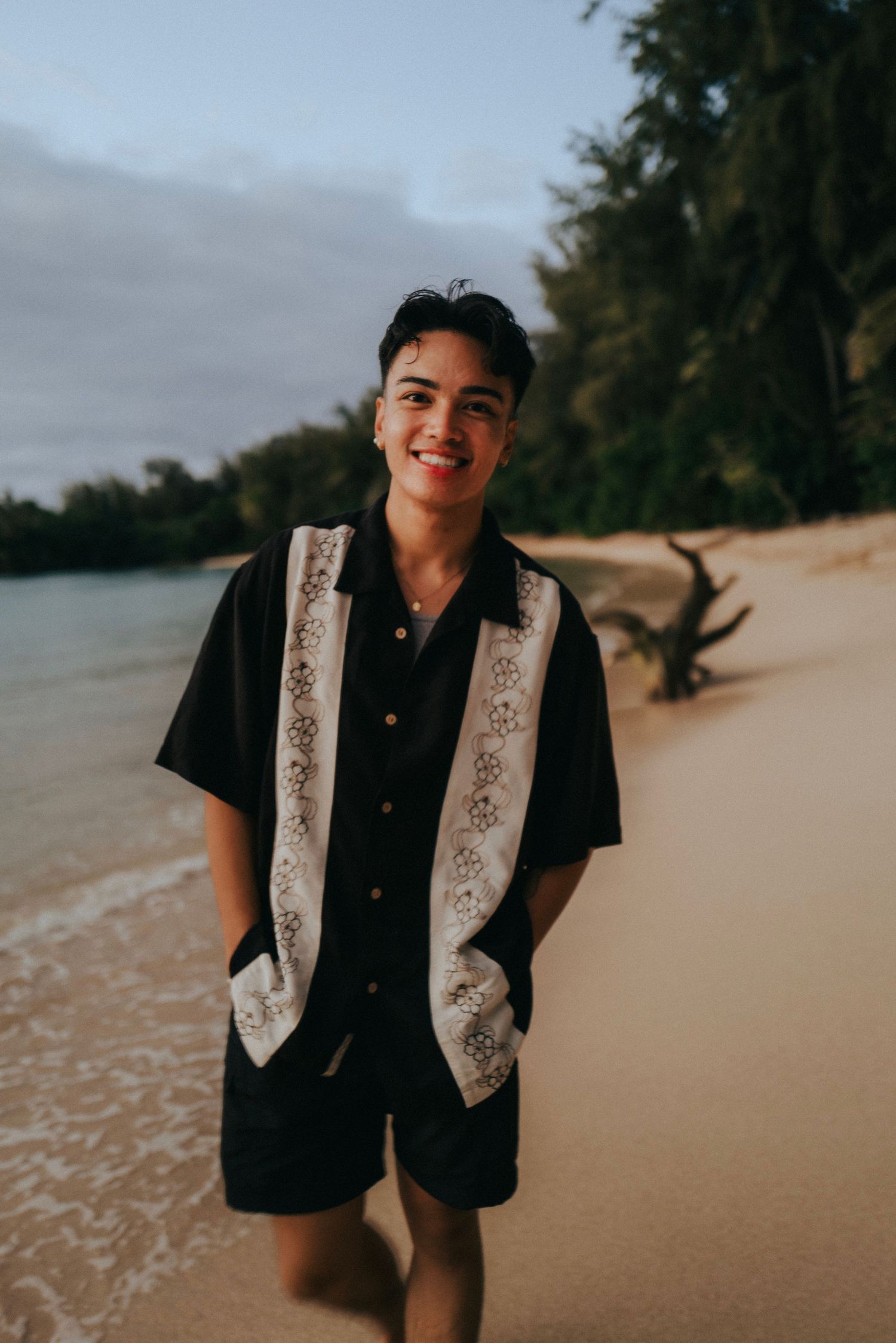 A person wearing a black and white patterned vest stands smiling on a tropical beach at sunset.