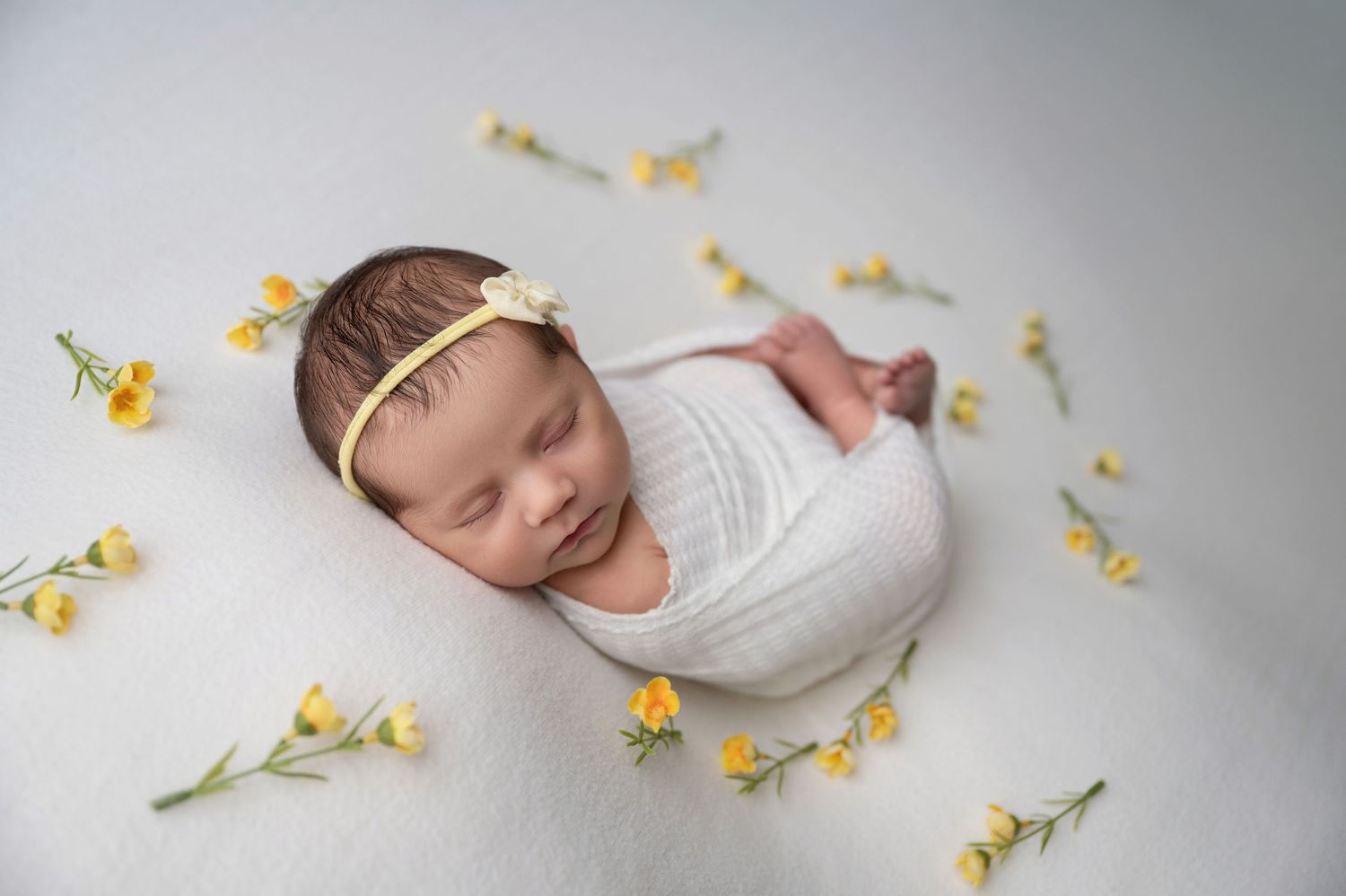 A sleeping newborn swaddled in white fabric lies on a blanket surrounded by delicate yellow flowers. Fresno Newborn Photographer