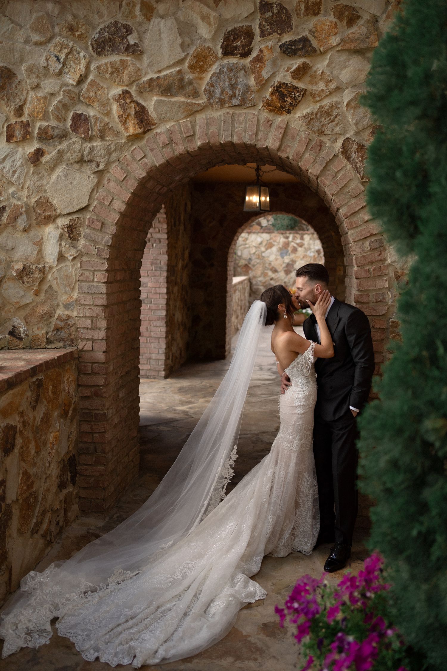 Wedding couple embraces under rustic stone archway with flowing veil and pink flowers.