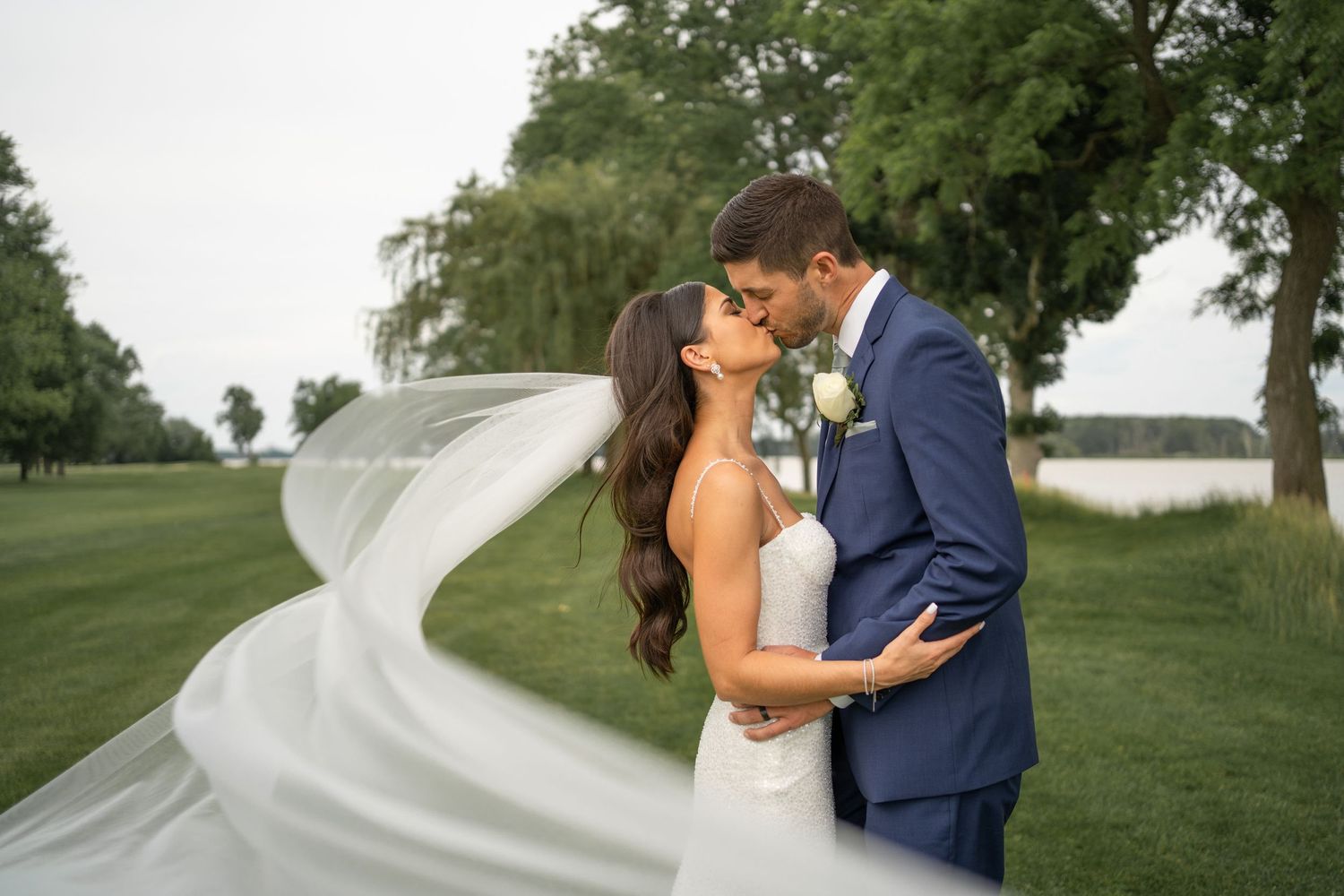 A flowing veil creates drama as newlyweds share a kiss by the waterfront.