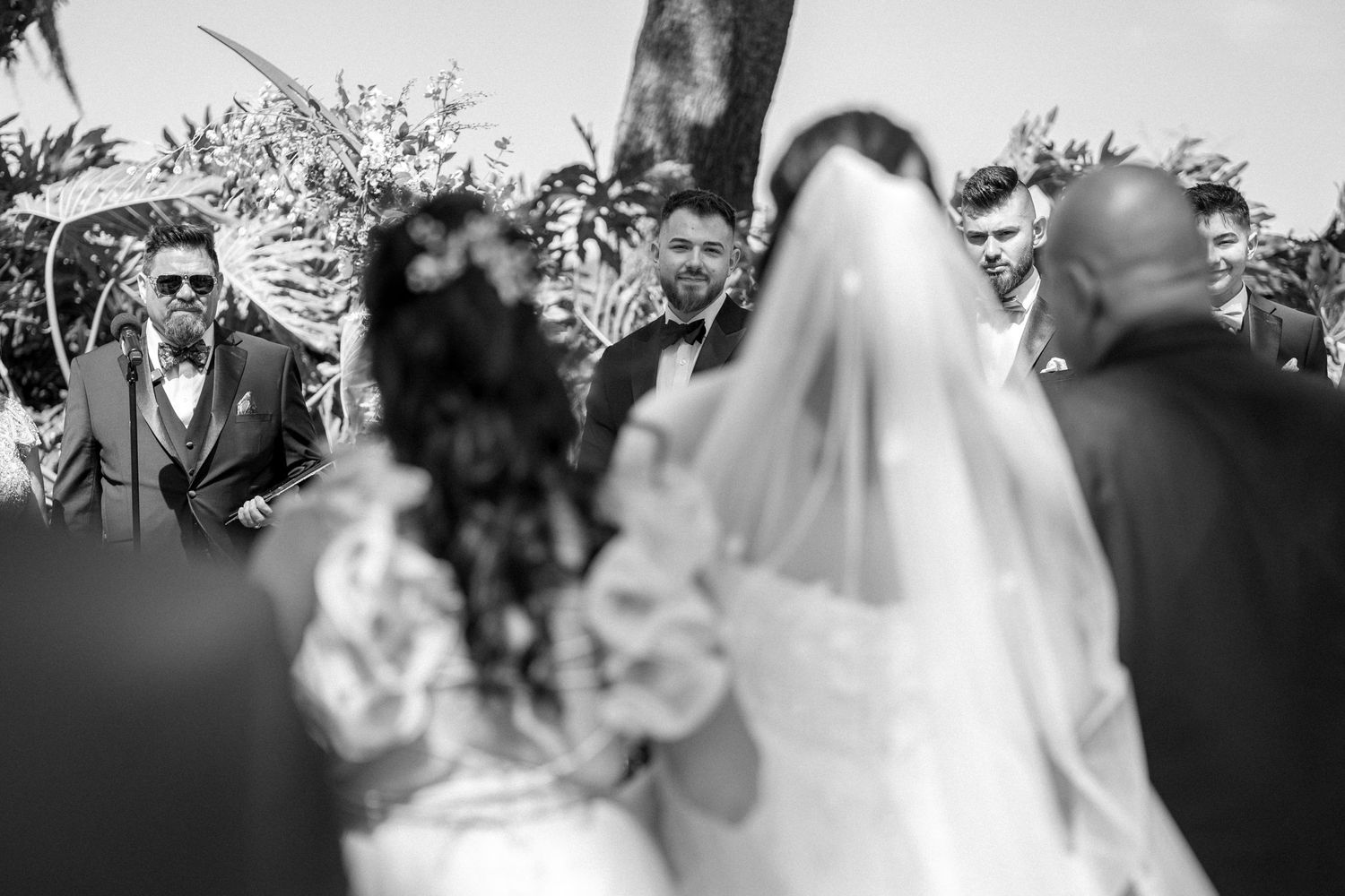 A wedding ceremony crowd gathers around the couple outdoors in black and white.