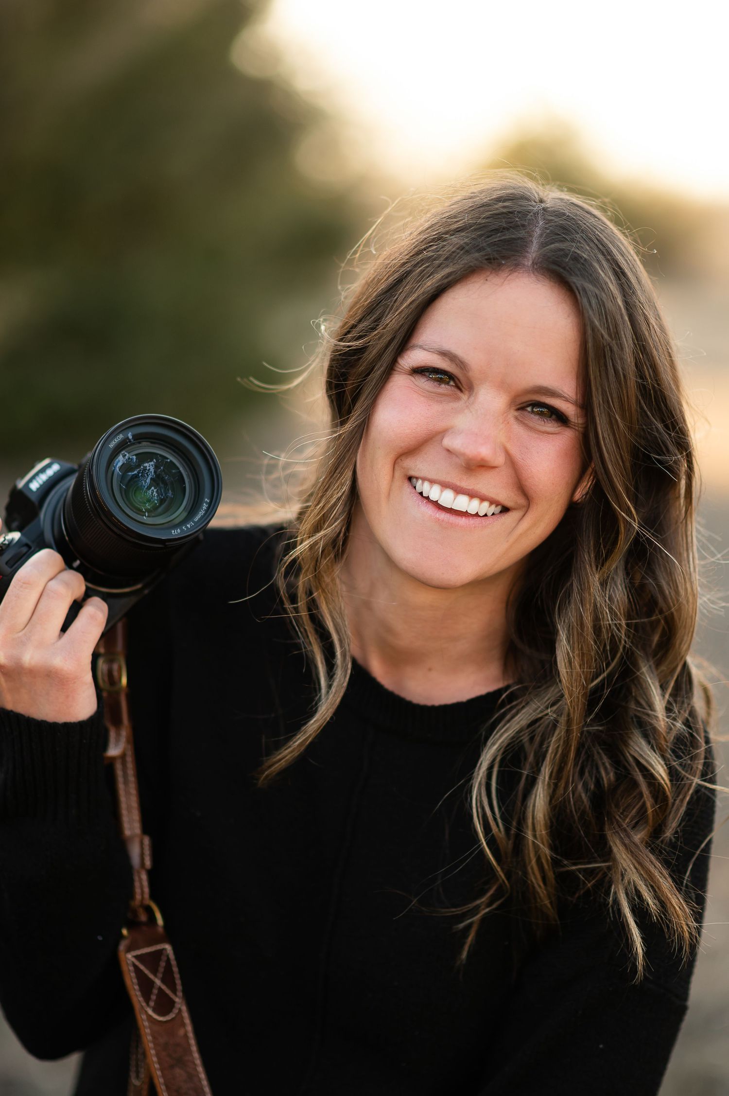 A headshot of a smiling female photographer holding a professional camera, looking at the camera with a friendly expression.