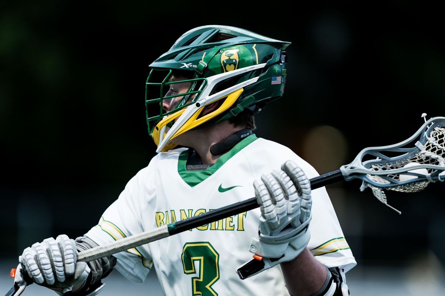 Lacrosse player in green and white uniform cradles ball while running downfield during competitive game action.