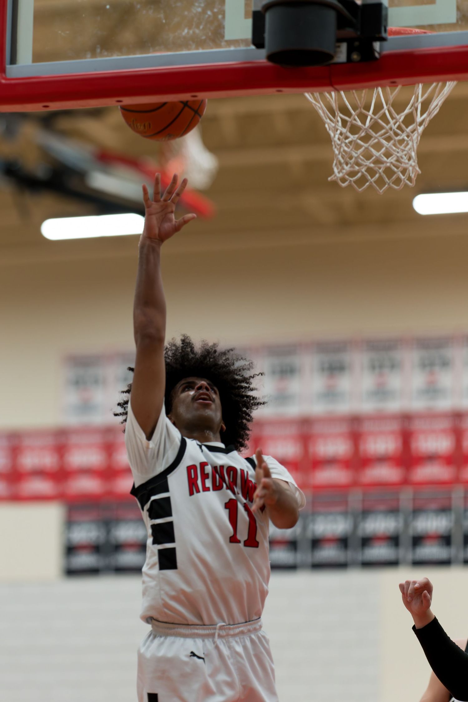 Basketball player in white jersey number 11 leaps toward the hoop during an indoor game against black-uniformed opponent.