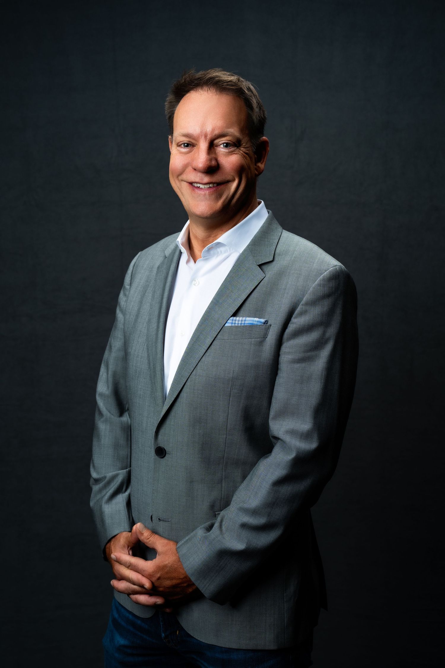 Professional studio portrait of someone wearing a gray suit jacket and white shirt against a dark background.
