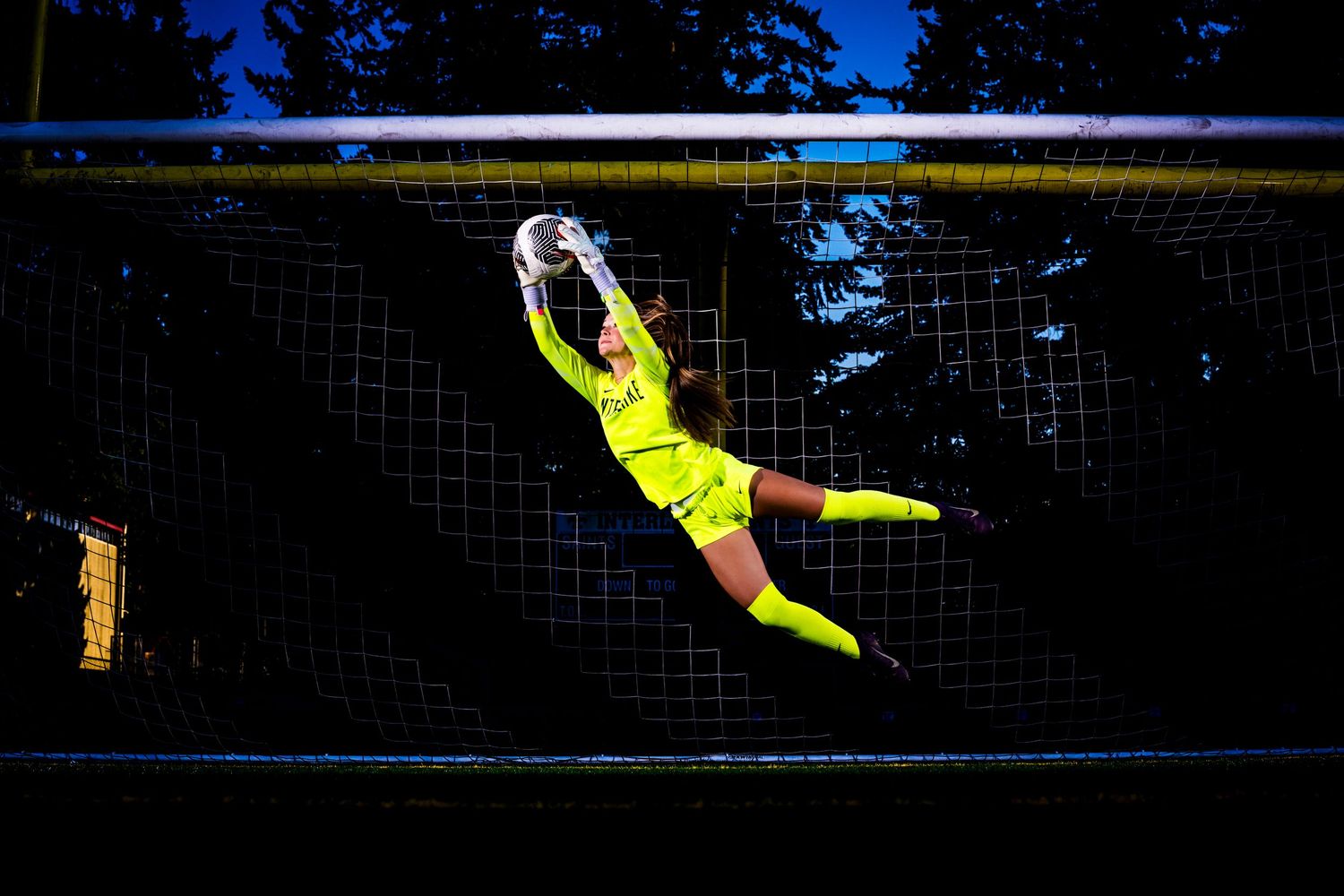 Soccer goalkeeper in bright yellow uniform makes dramatic diving save attempt during night match.