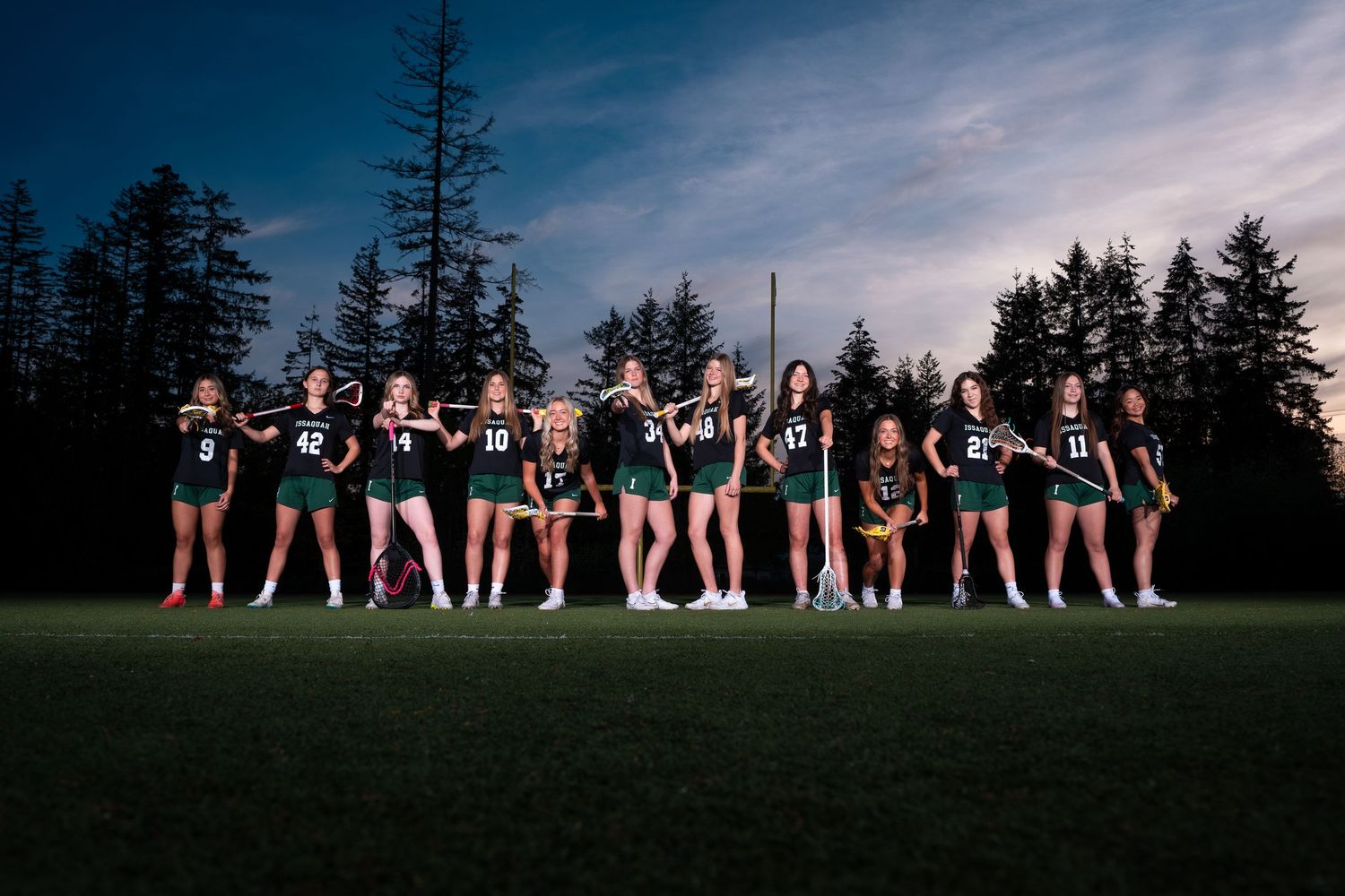 A sports team poses together on a grass field at dusk with evergreen trees silhouetted against the darkening sky.