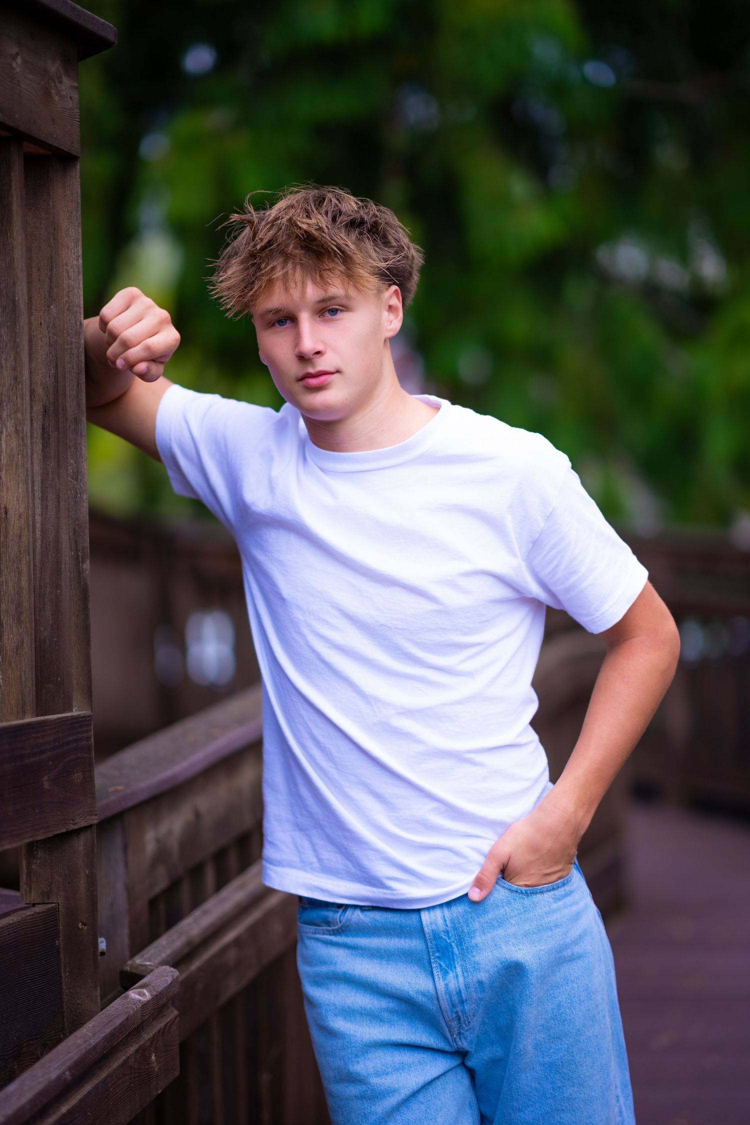 A person in a white t-shirt and blue jeans poses casually against a wooden railing in an outdoor setting.