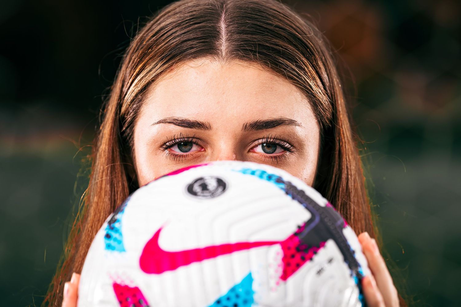 Close-up shot of a Nike Premier League soccer ball with blue and pink accents held up against a blurred background.