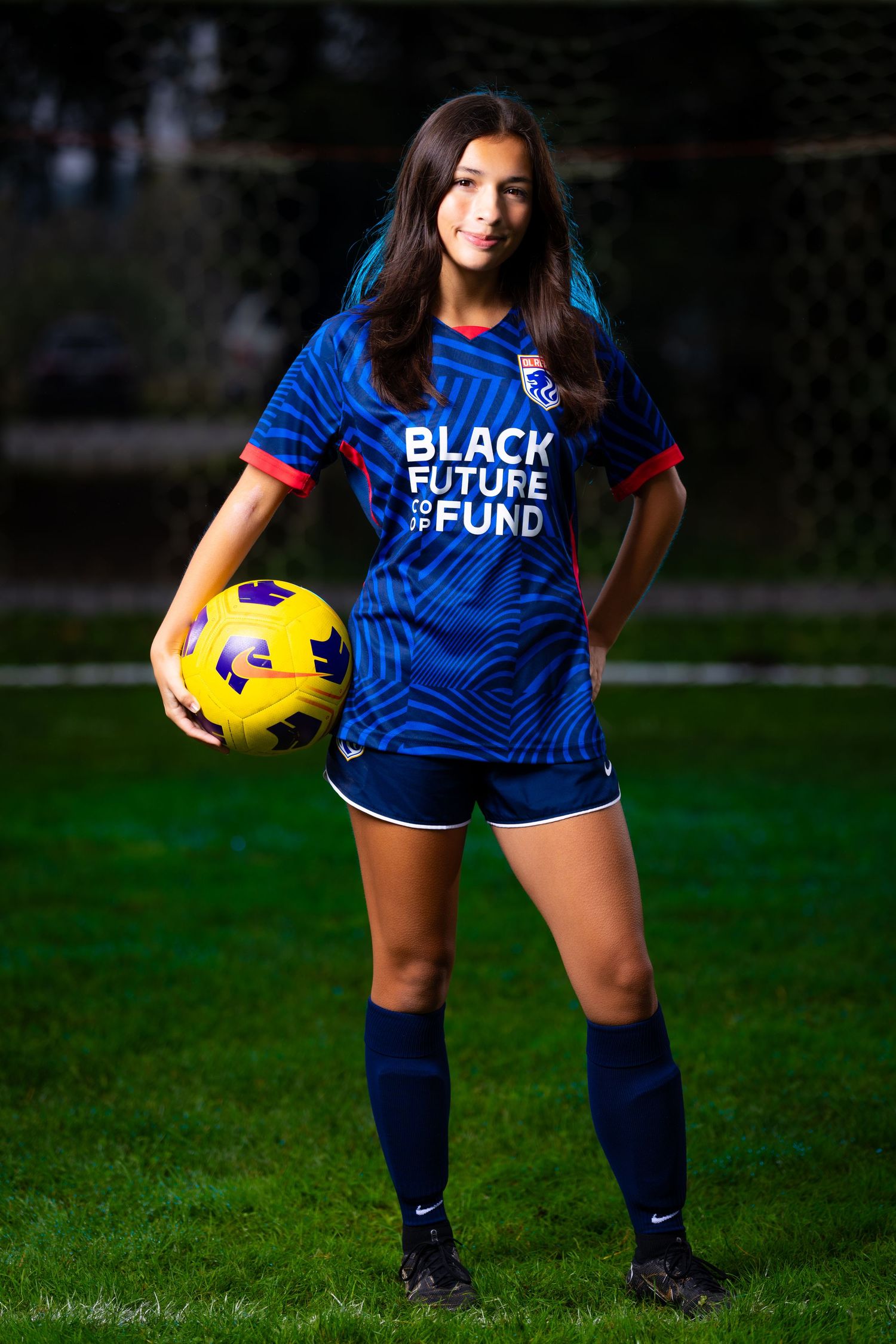 Soccer player in blue Black Future Fund jersey holds yellow ball while posing on grass field at dusk.