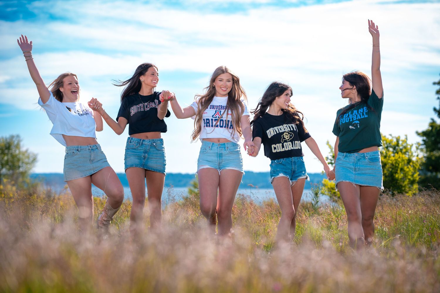 A group of friends in casual summer outfits jump joyfully together in a sunny meadow with blue sky in the background.