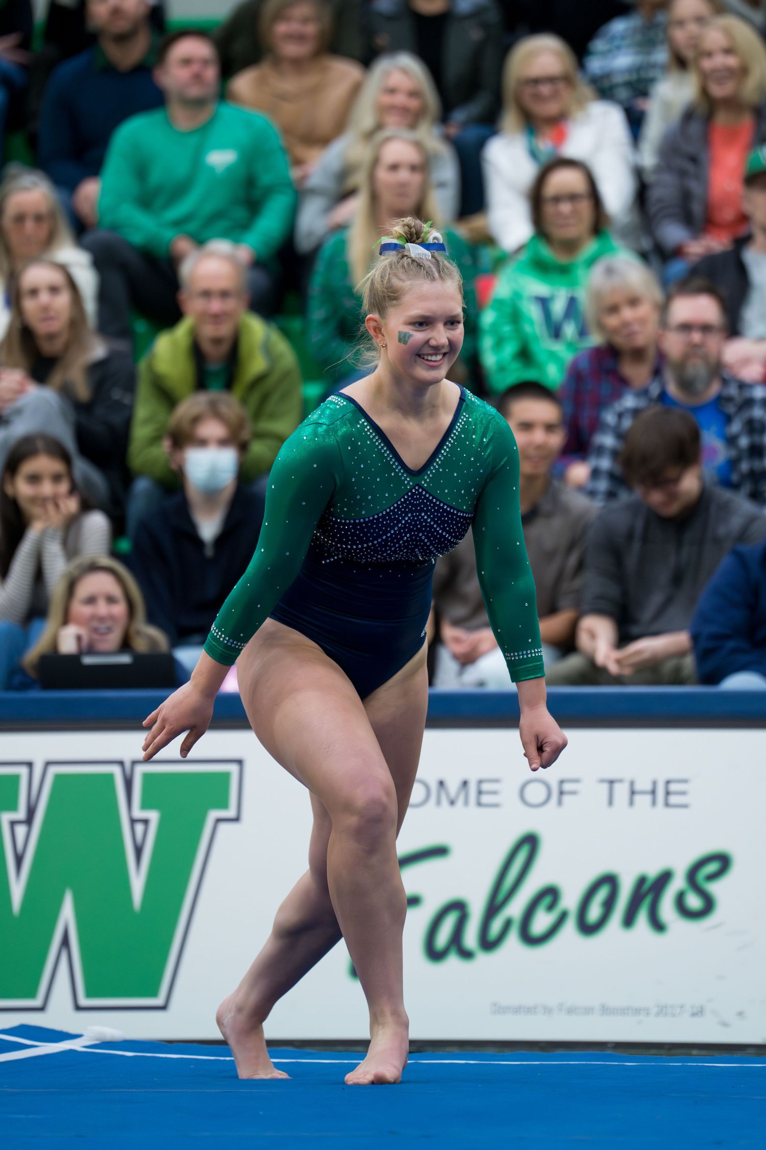Gymnast in green and navy leotard performs floor routine at competition with crowd in background.