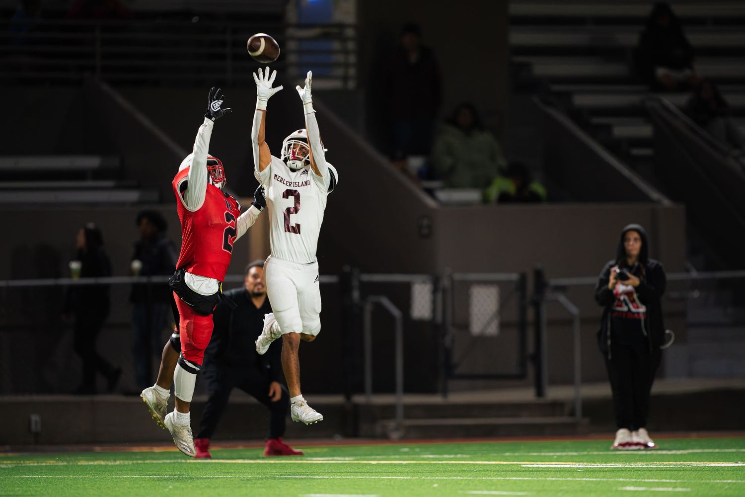 Two football players leap high in the air competing for a catch during a nighttime game under stadium lights.