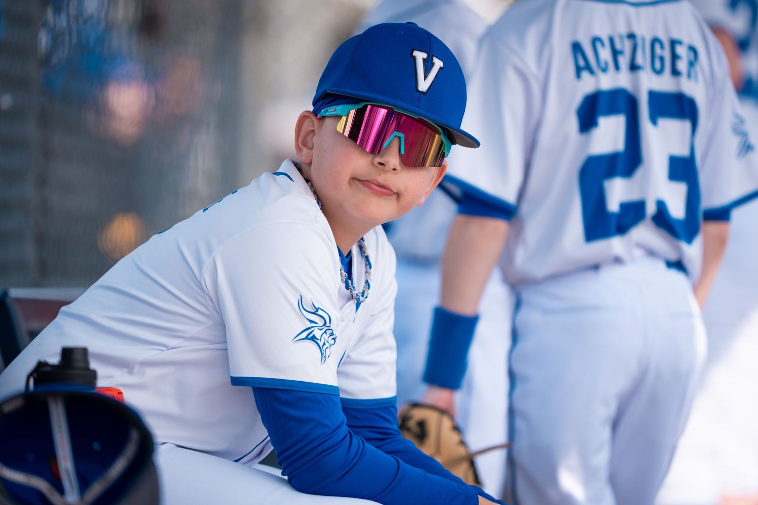 Baseball players in white and blue uniforms sit in the dugout during a game at a stadium.