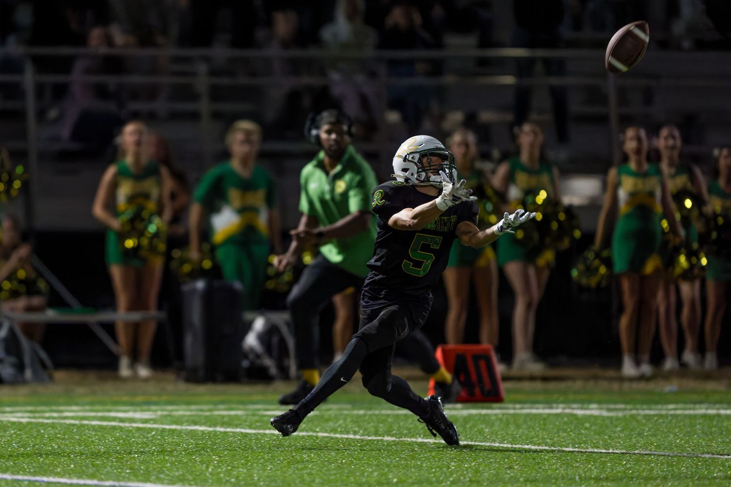 Football player kicks field goal attempt during nighttime game with crowd watching from stadium stands in background.