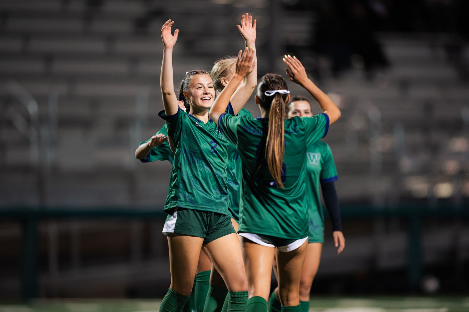 Soccer players in green uniforms celebrate a goal with raised arms and joyful expressions during a match.