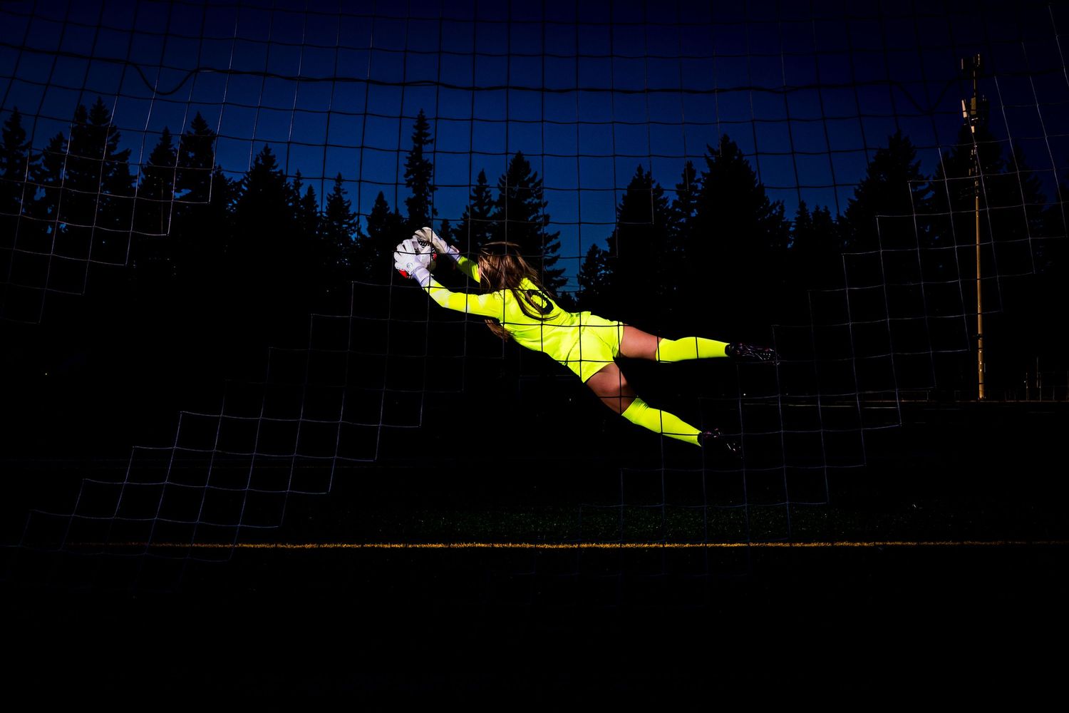 A soccer goalkeeper in bright yellow uniform makes a diving save during a night match under stadium lights.
