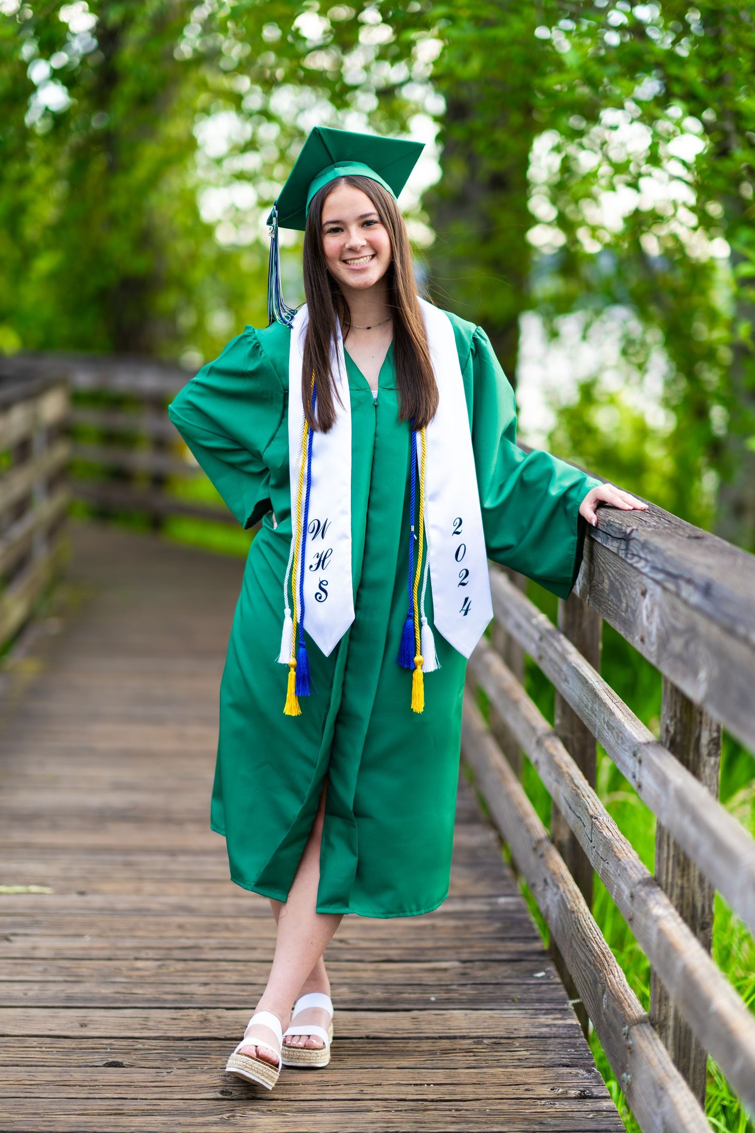 A graduate in emerald green cap and gown poses on a wooden bridge surrounded by lush greenery.