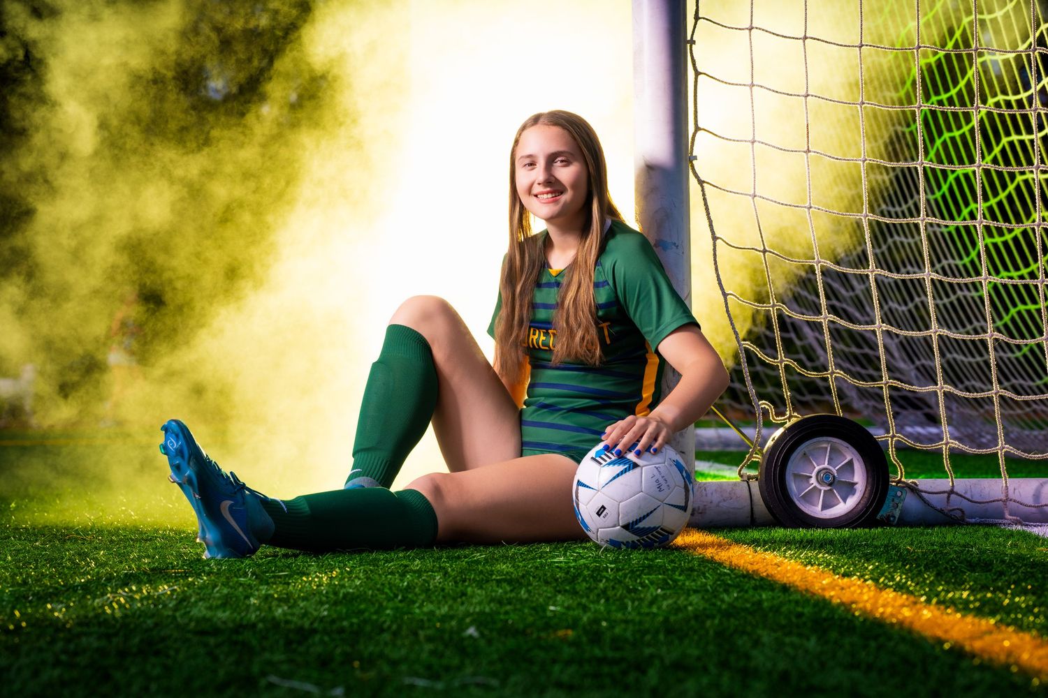 Soccer player in green uniform sitting by goal net with soccer ball on sunny field.