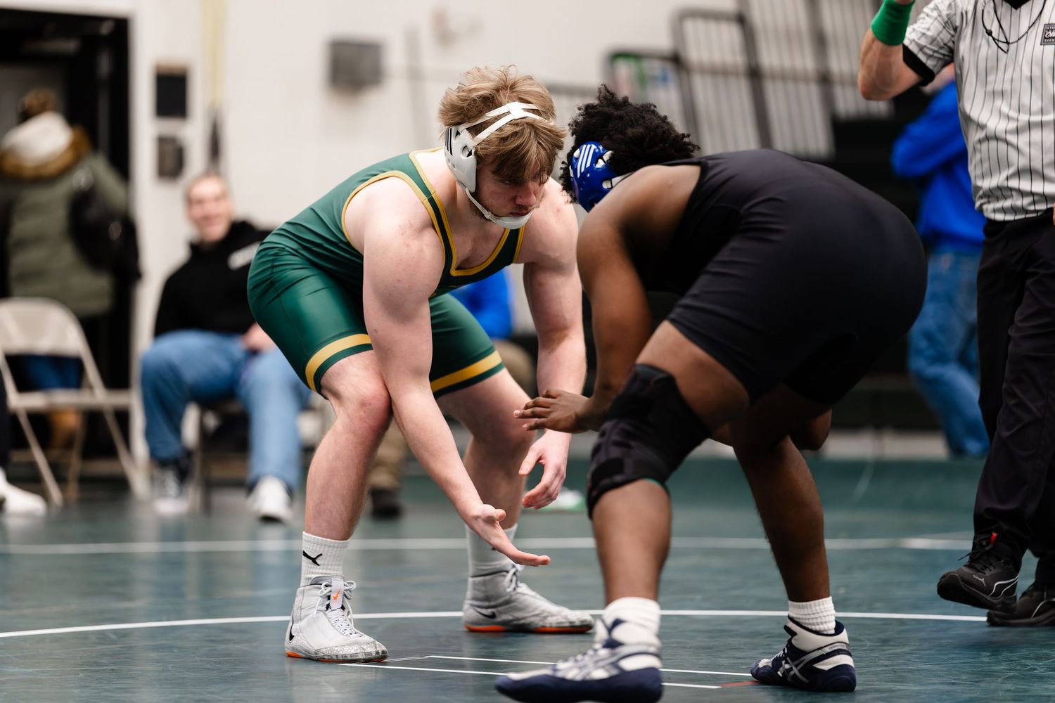 Two wrestlers in green and black uniforms face off in starting position during a competitive match on the mat.