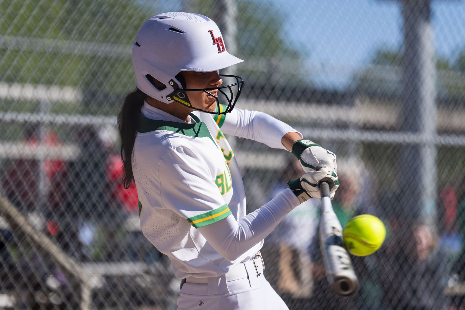 A softball player in a white uniform swings the bat at a bright yellow ball during a game.