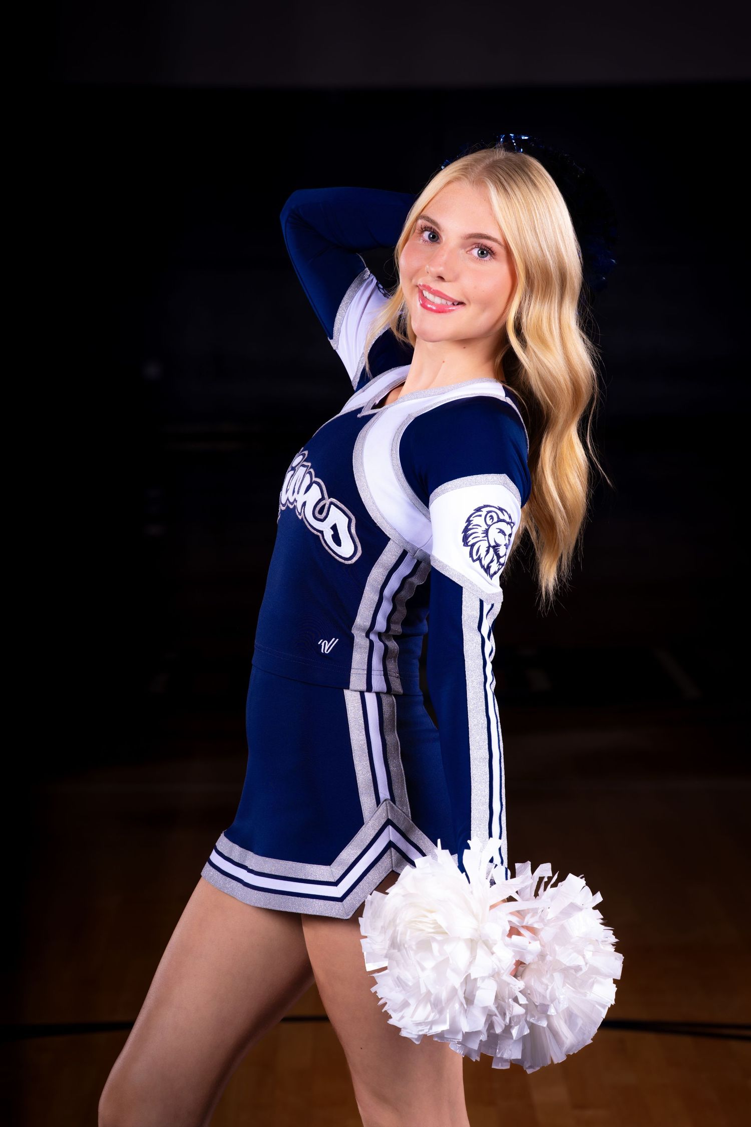 Cheerleader in navy and white uniform poses with pom pom against dark background.