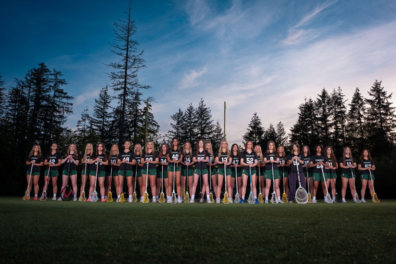 A large group of cheerleaders in red and white uniforms pose in formation on a grassy field at dusk with evergreen trees behind them.