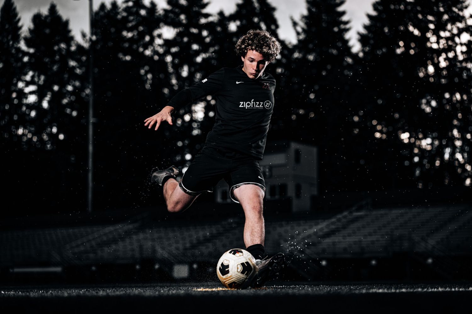 Soccer player in black uniform kicks ball during nighttime practice session on dark field with evergreen trees in background.