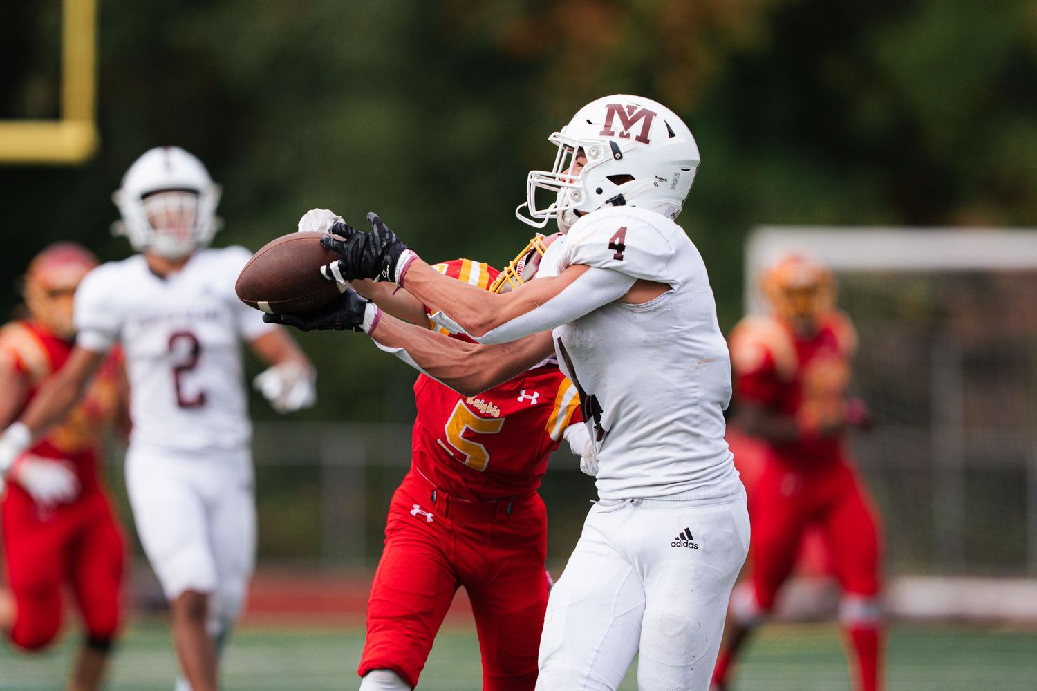 A high school football game shows players in red and white uniforms competing on the field during a play.