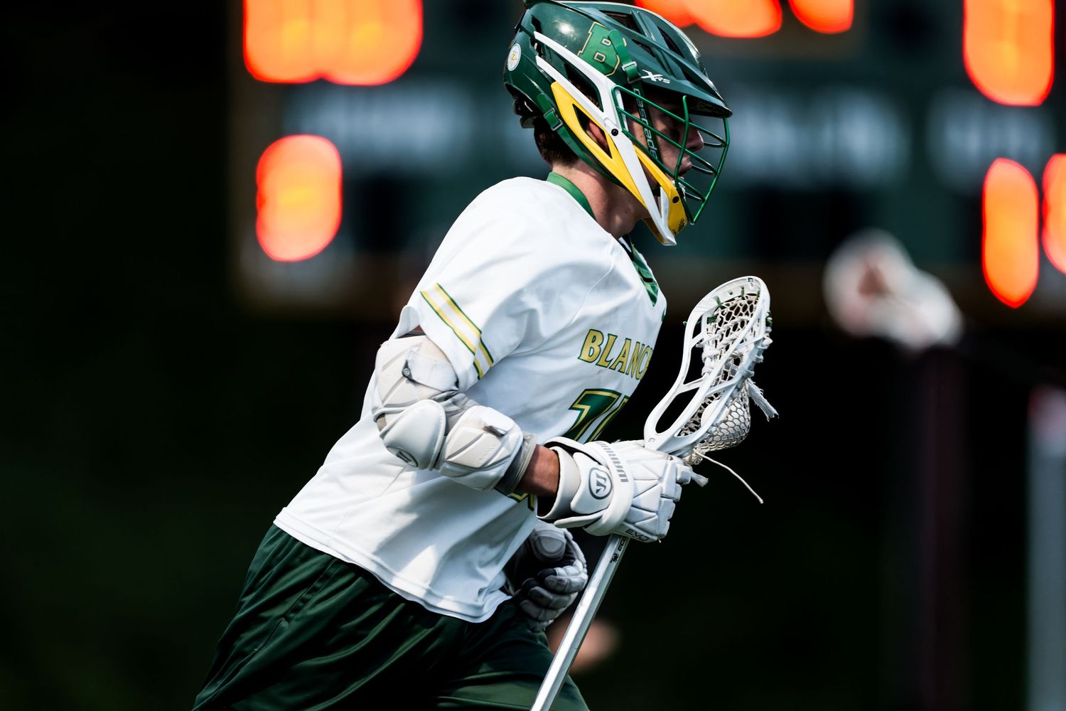 Lacrosse player in white jersey and green helmet holds stick during night game with orange lights in background.