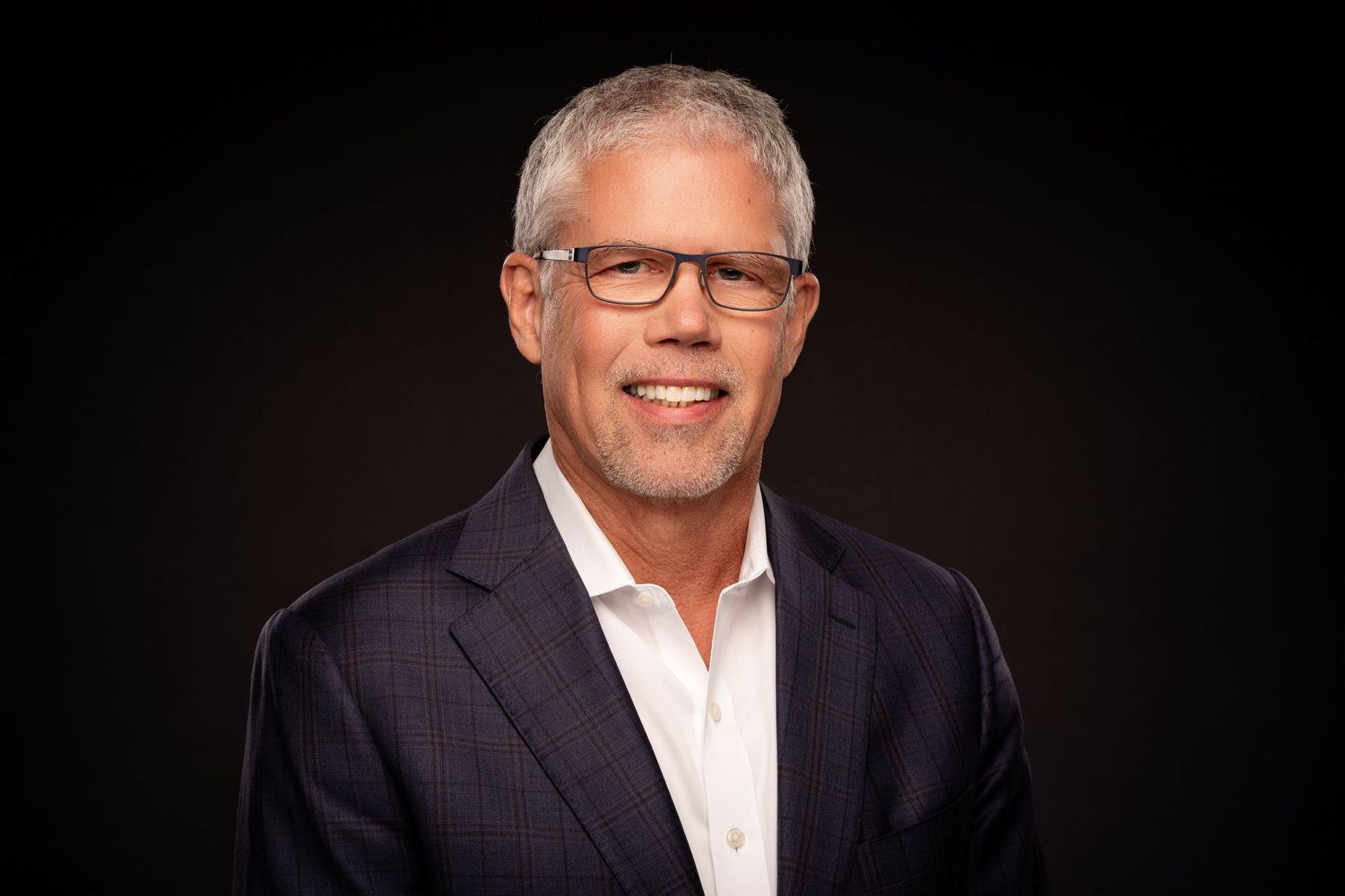 Professional headshot portrait of person in glasses and white shirt smiling against dark background.