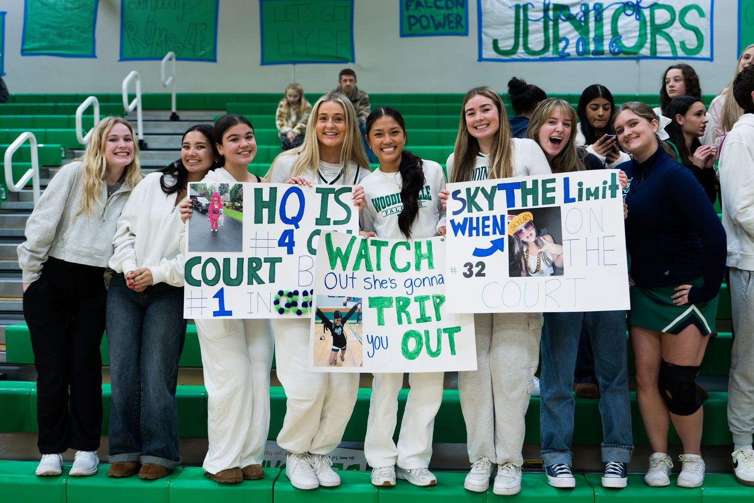 Students hold up supportive signs in green school colors while standing along a gymnasium wall during a sporting event.