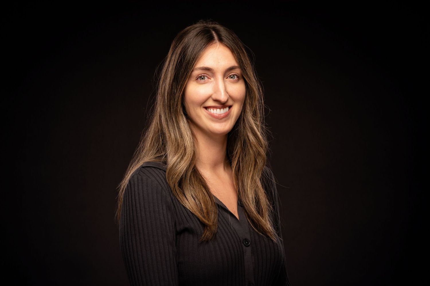 Professional headshot portrait of someone with long brown hair wearing a black pinstripe top against a dark background.
