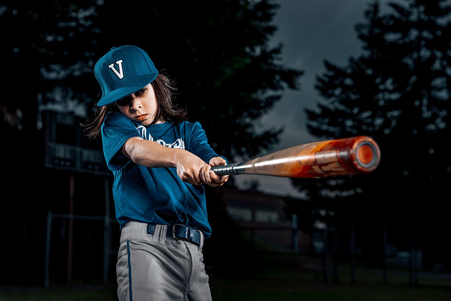 Youth baseball player in blue uniform swings orange bat during evening practice session on field.