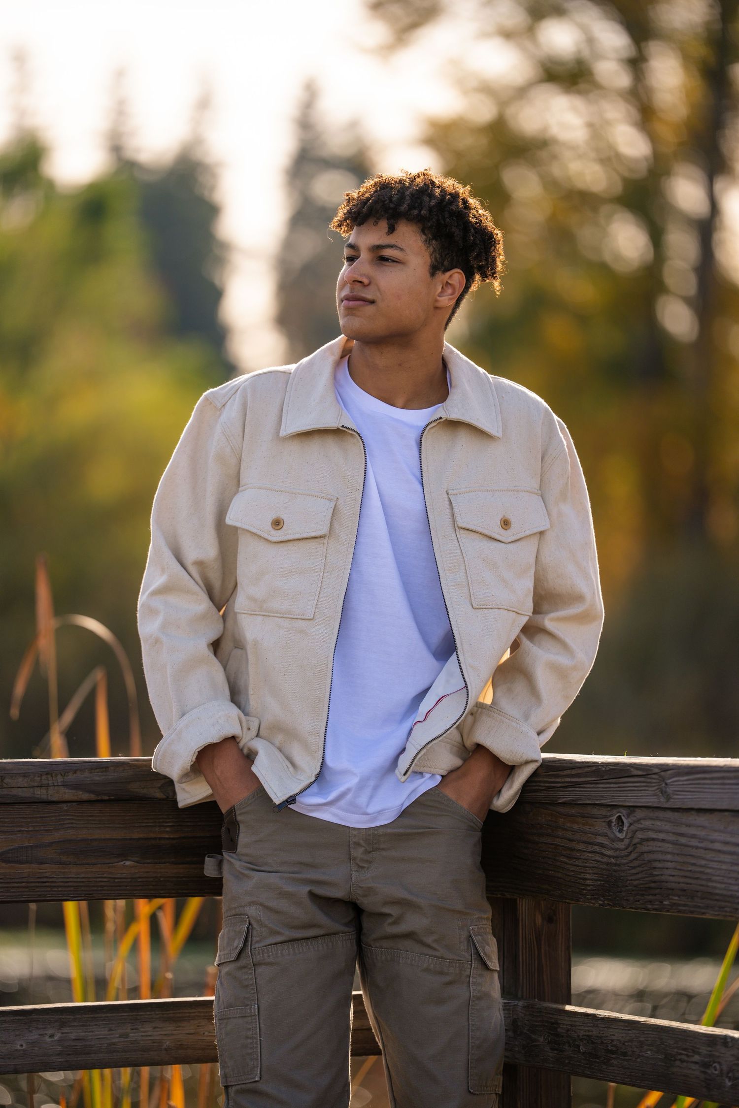 Person in beige jacket and white t-shirt leans against wooden fence during golden hour sunset.