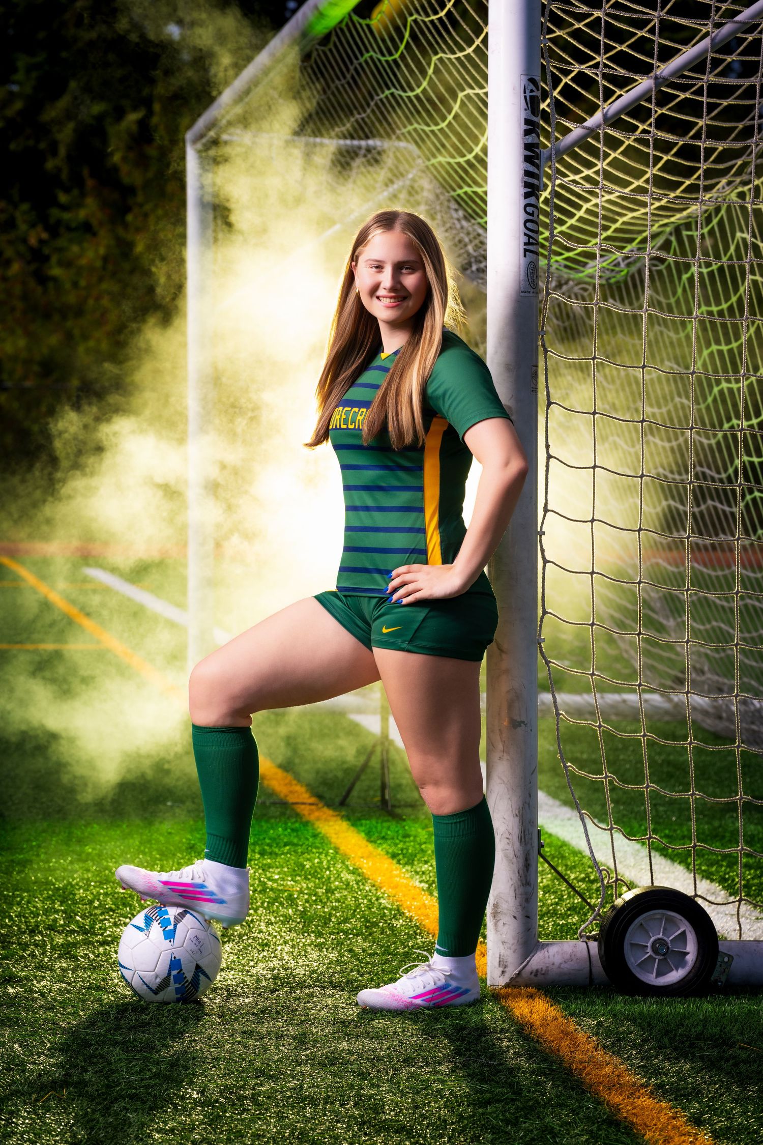Soccer player in green uniform poses with ball and goal net in atmospheric lighting.