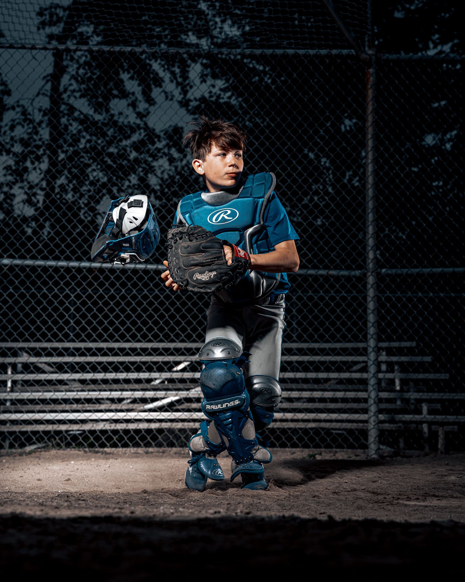 Youth catcher in full blue gear poses in dramatic lighting against chain link fence.