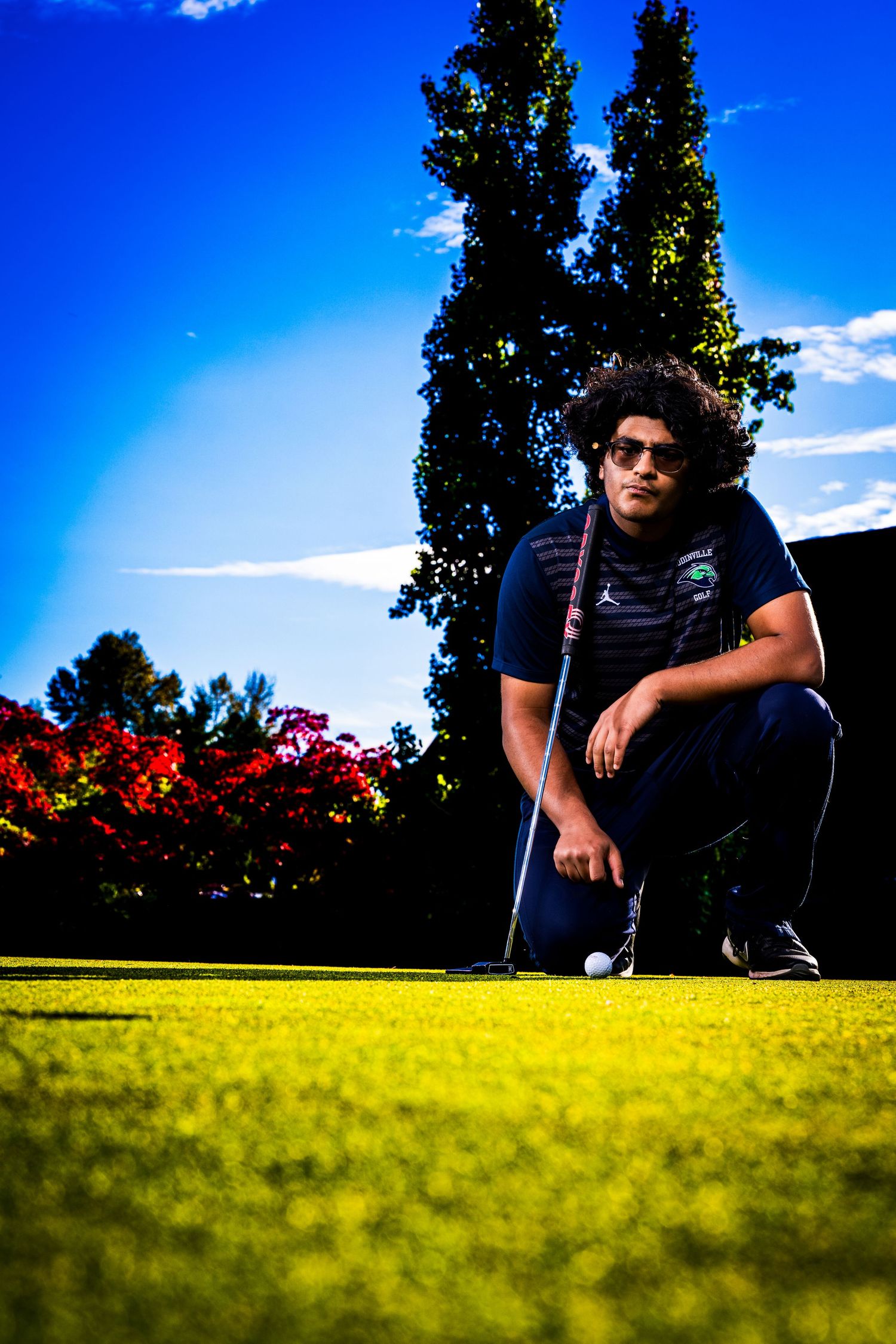 Golfer crouches to analyze putting green against vibrant blue sky and flowers.