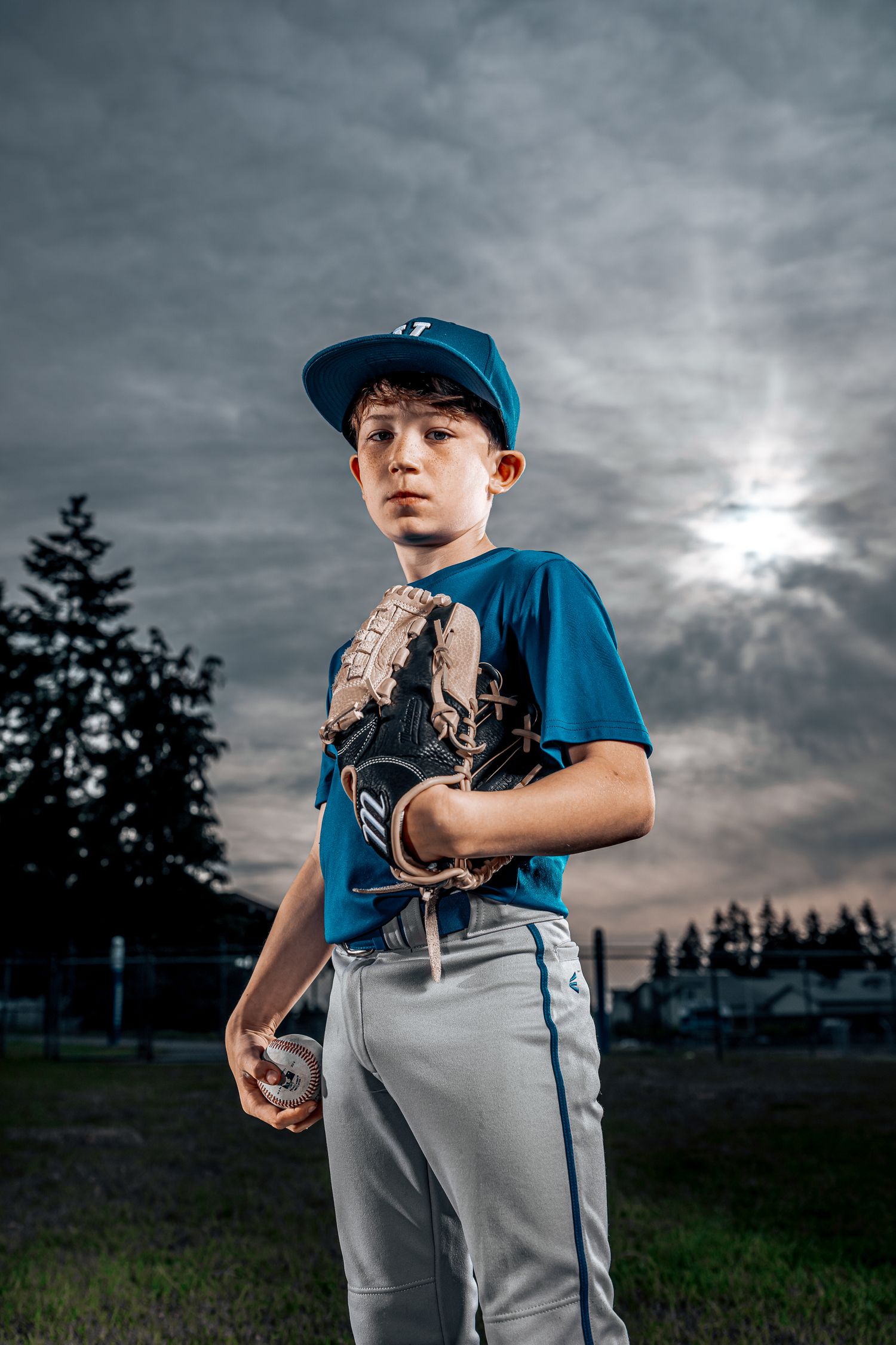 Dramatic portrait of youth baseball player in blue uniform against moody sunset sky.