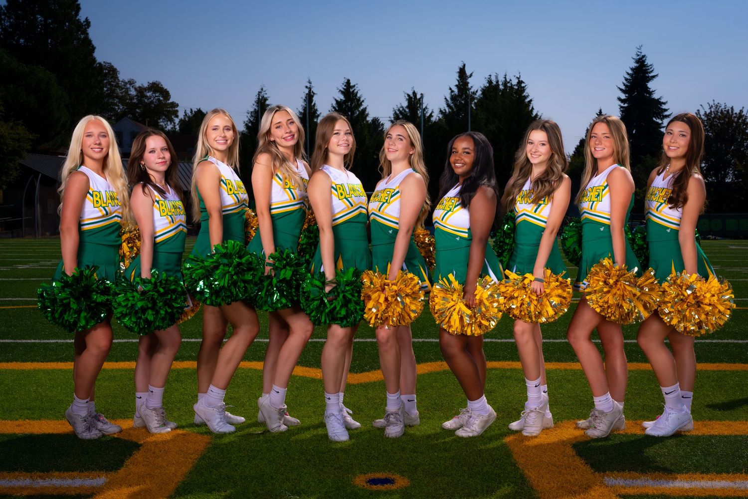 Cheerleaders in green uniforms and white shoes line up with yellow pom poms on a sports field at sunset.