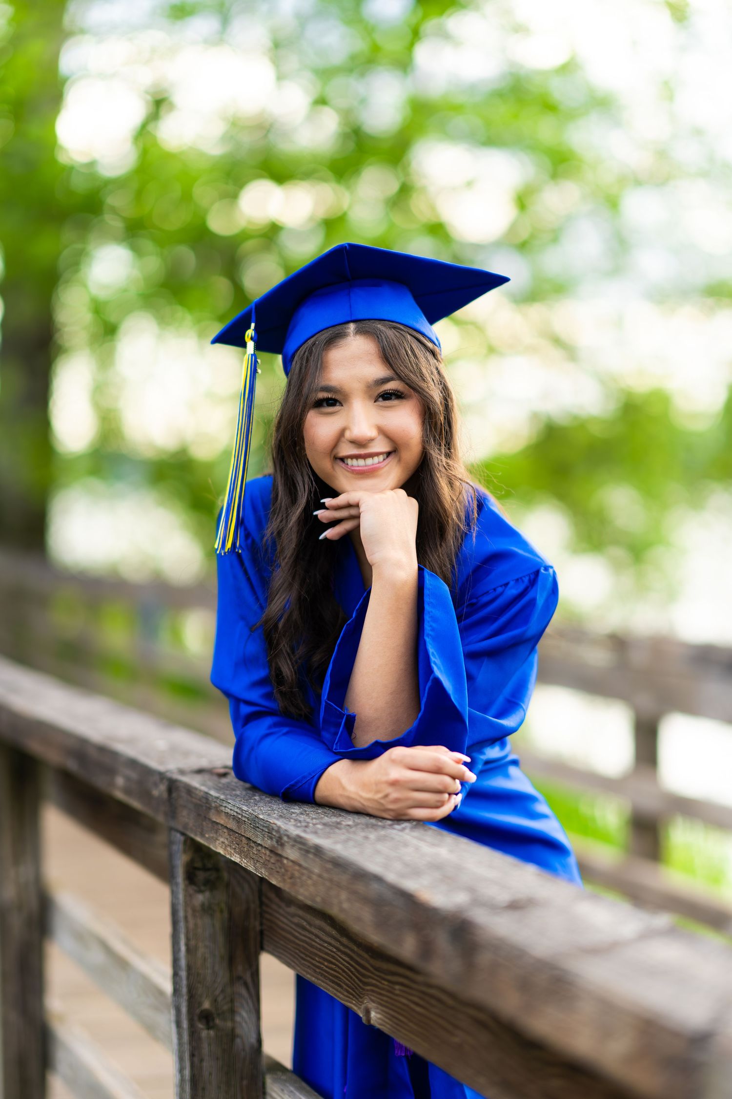 A graduate in royal blue cap and gown poses playfully on wooden bleachers surrounded by green trees.