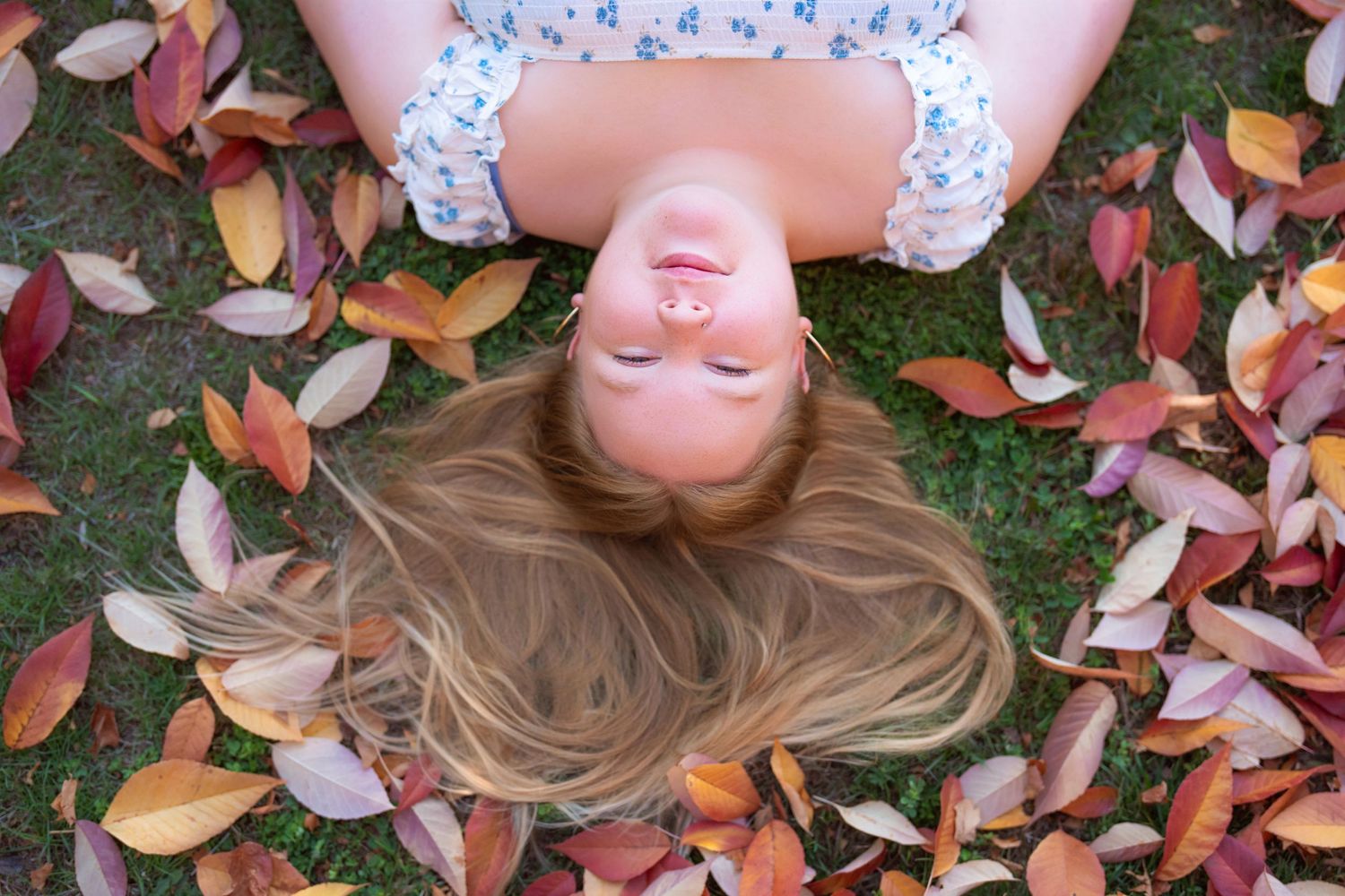 A person lying in colorful autumn leaves on grass while wearing a pink sweater and smiling at the camera.