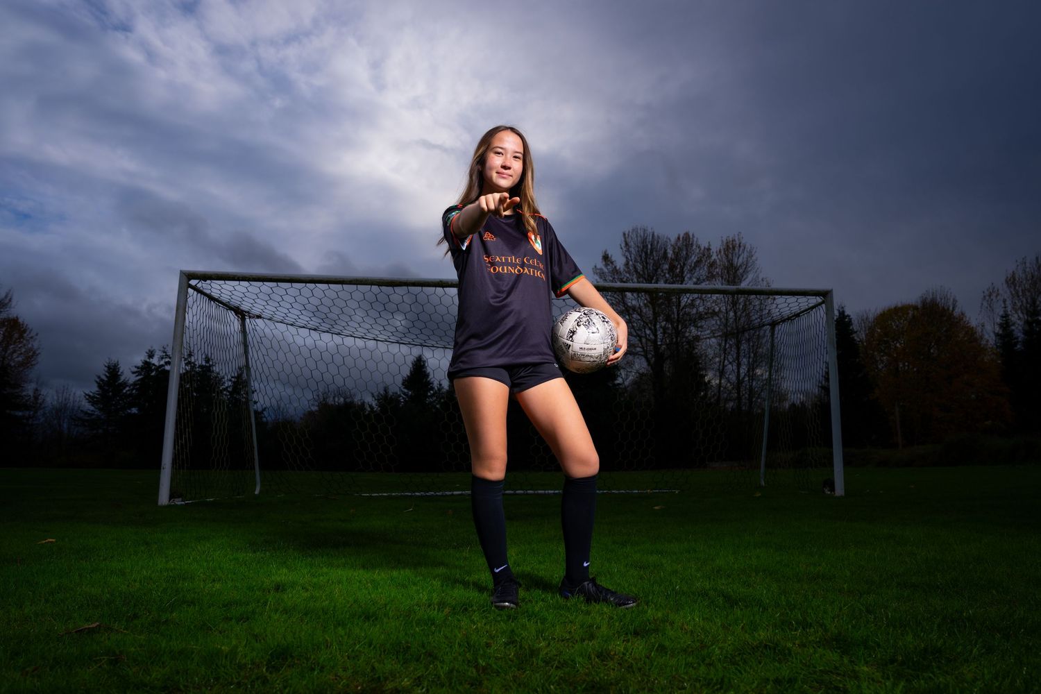 Soccer player poses with ball on field at dusk with dramatic cloudy sky and goal in background.