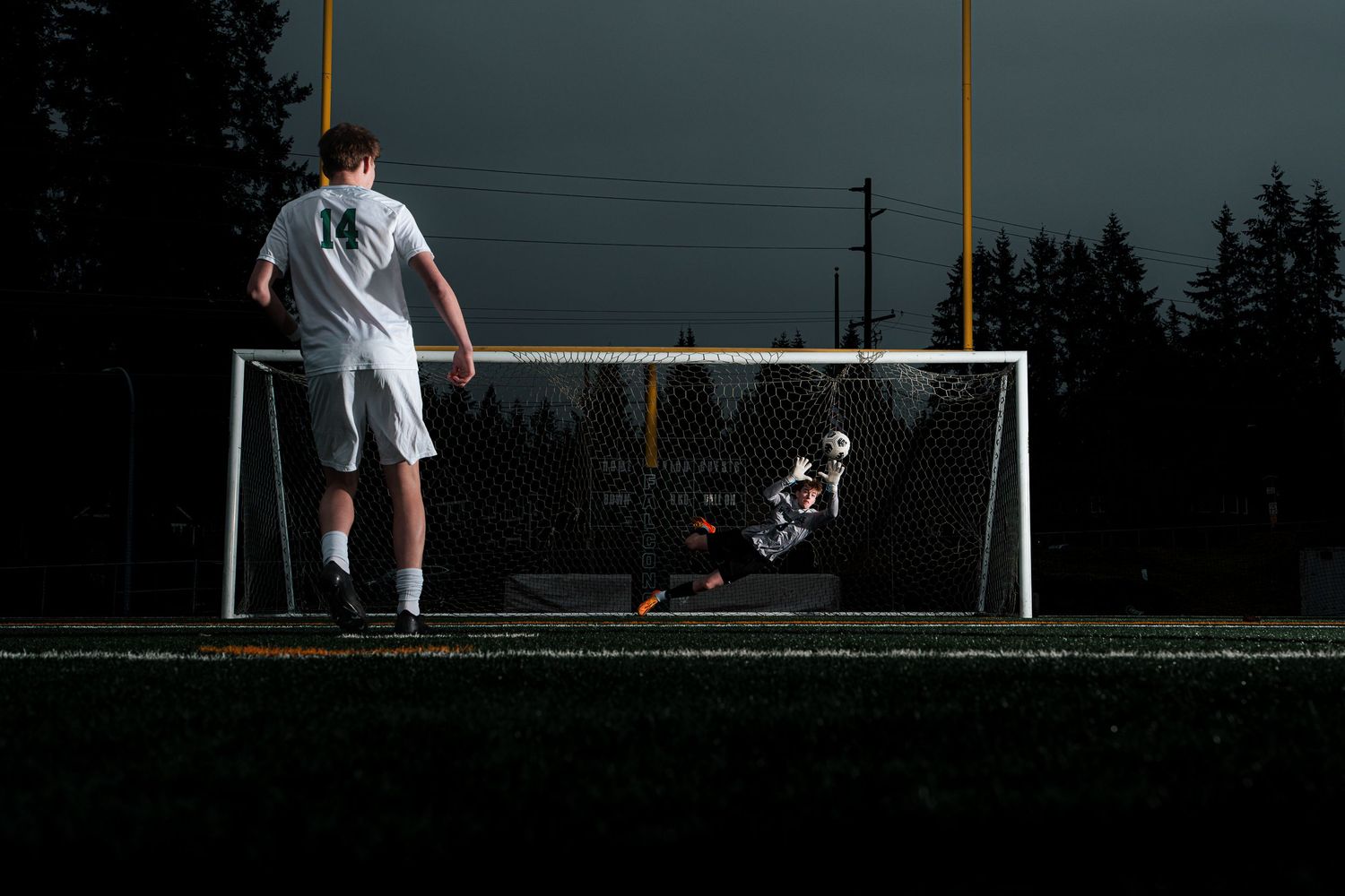 Soccer goalkeeper in white uniform prepares to defend goal during match on outdoor field at dusk.