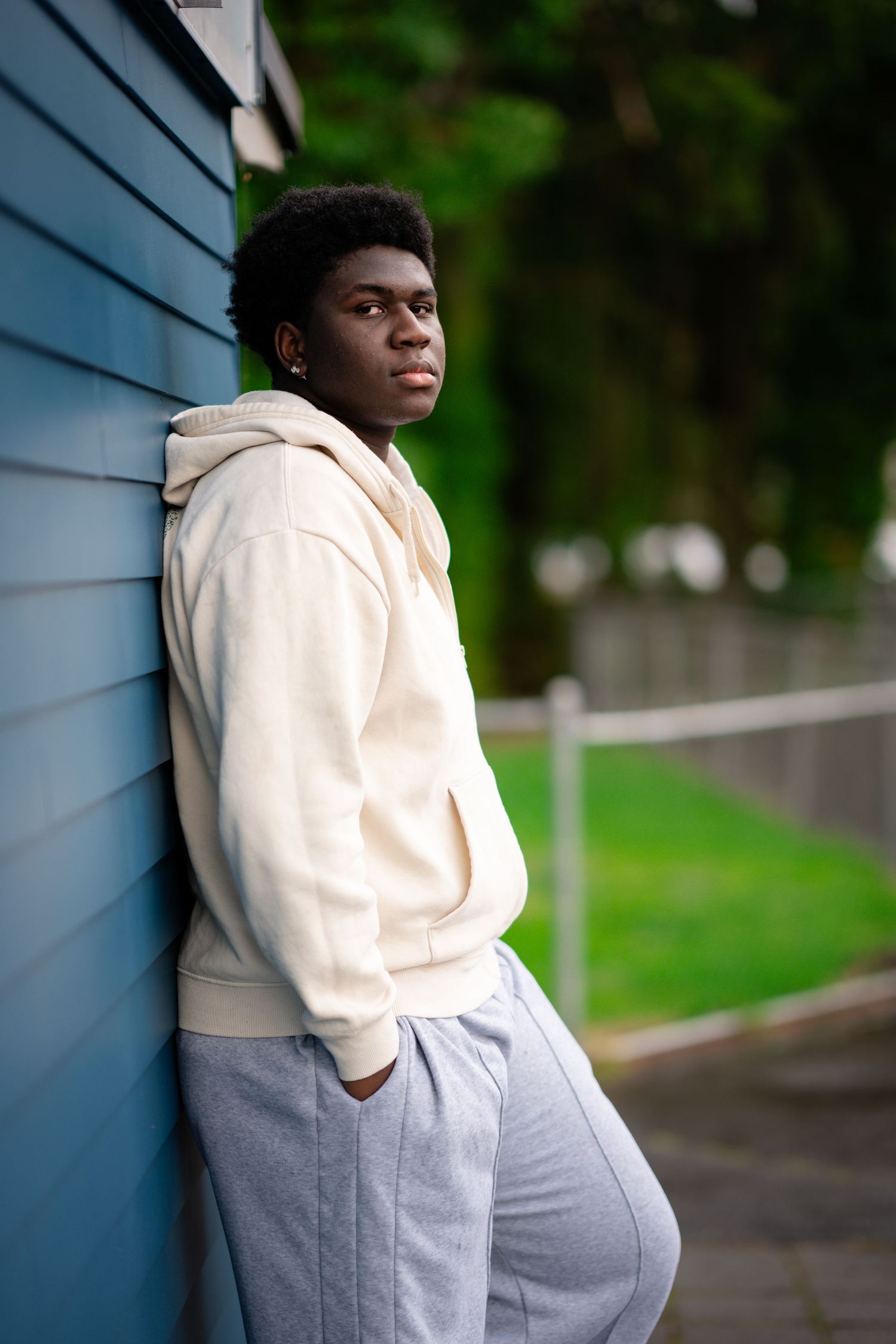 Person in cream hoodie and gray pants poses against blue wall in outdoor setting.